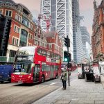 Red double-decker bus passing through central London street with historic and modern buildings.