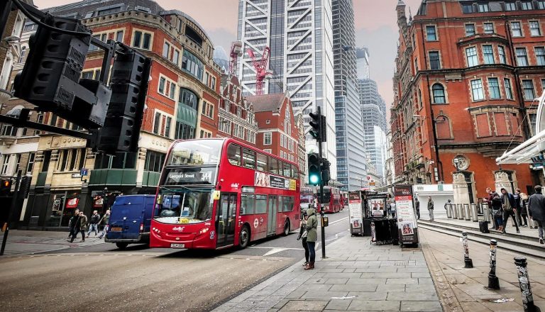 Red double-decker bus passing through central London street with historic and modern buildings.