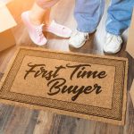 Doormat with “First Time Buyer” beside moving boxes and two people’s feet