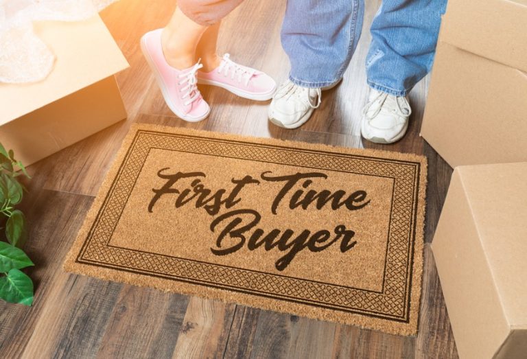 Doormat with “First Time Buyer” beside moving boxes and two people’s feet