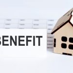 Wooden house model beside a paper with the word “BENEFIT,” symbolizing housing benefit payments.