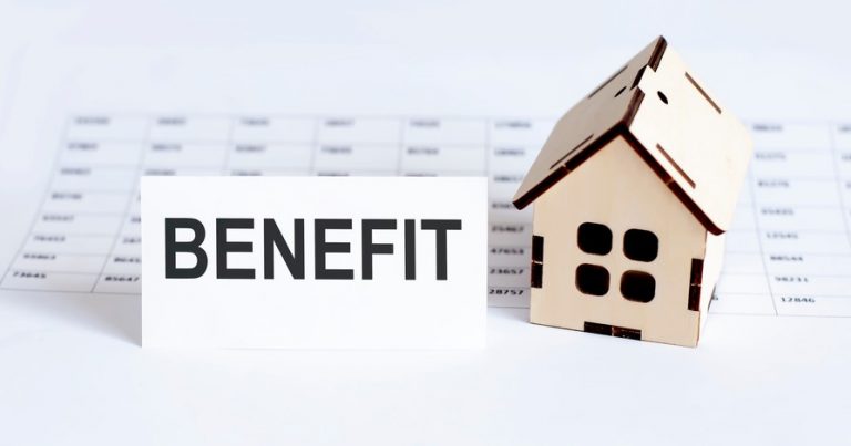 Wooden house model beside a paper with the word “BENEFIT,” symbolizing housing benefit payments.