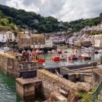 Colourful fishing boats moored in a Cornish harbour surrounded by stone cottages and green hills.