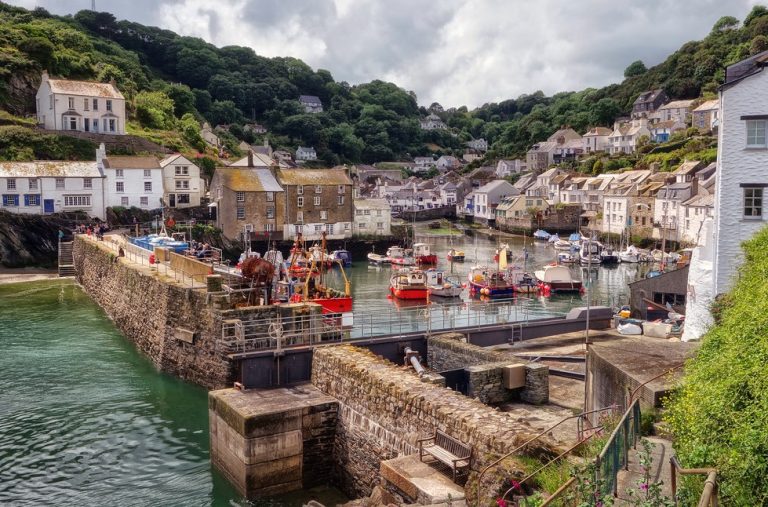 Colourful fishing boats moored in a Cornish harbour surrounded by stone cottages and green hills.