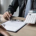 A man in a suit holding a calculator, a clipboard, a small model house at a desk sitting opposit a person,