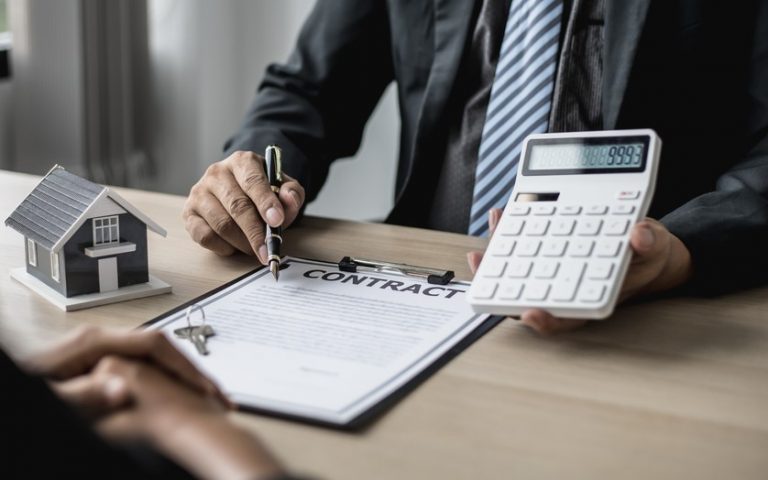 A man in a suit holding a calculator, a clipboard, a small model house at a desk sitting opposit a person,