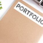 Brown folder labeled “Portfolio” beside a pen, notebook, and plant on a white desk.
