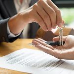 Hands handing over keys with renters agreement paperwork on a table