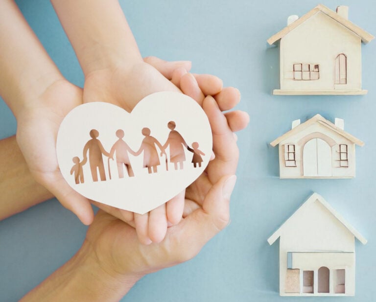 Hands holding a paper heart with a family silhouette beside miniature house models, symbolizing family wealth and legacy planning