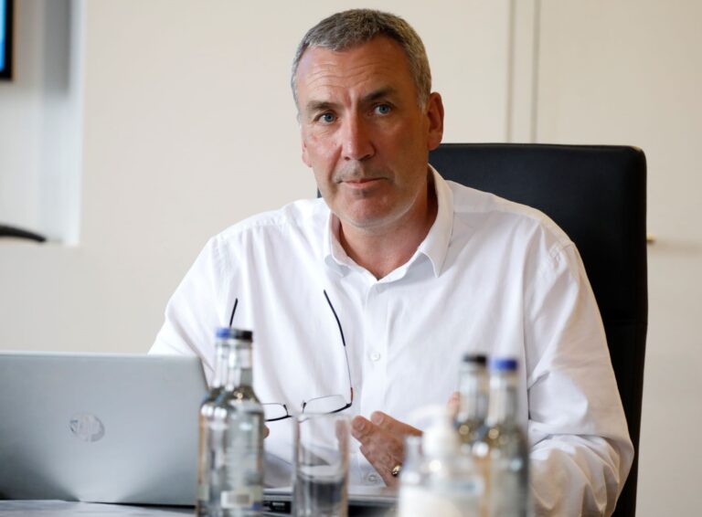 Man in white shirt speaking at meeting with laptop and water bottles on table
