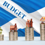 Stacked coins with model houses and Scottish flag illustrating budget impact on housing and landlords