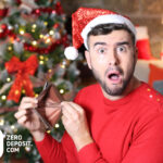Man in Santa hat shocked by empty wallet in front of Christmas tree