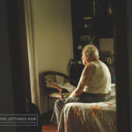 Elderly man sitting on bed looking out window, symbolizing vulnerable tenants facing housing barriers