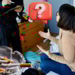 A lady sitting on a bedrrom floor looking upto another person who is writing on a clipbaord and a question mark