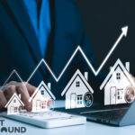 Small model houses with arrow index pointing upwards above the houses, a person sitting in front of a laptop and a calculaor at a desk