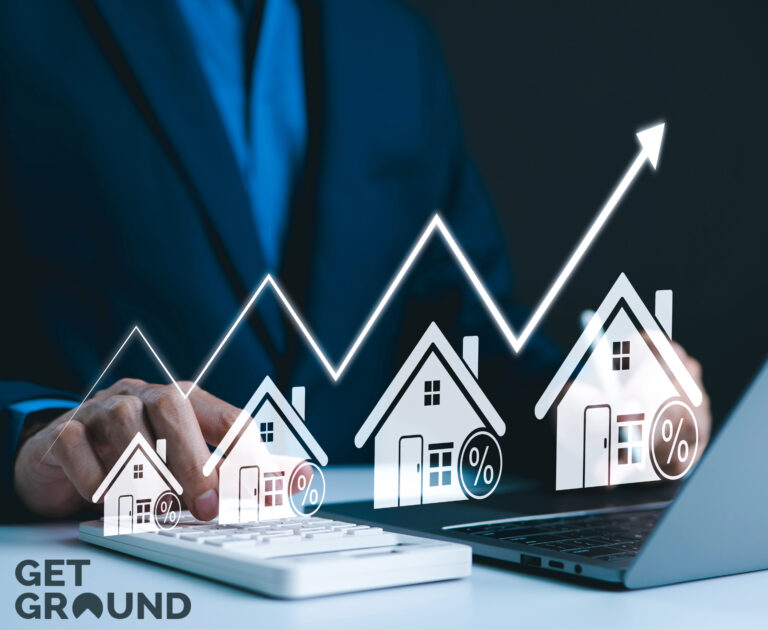 Small model houses with arrow index pointing upwards above the houses, a person sitting in front of a laptop and a calculaor at a desk