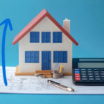 A model house, a blue arrow pointing upwards, a calculator, pen sitting on papers with a blue background
