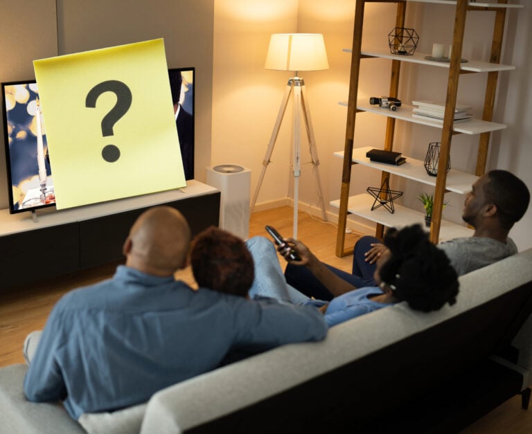 A family sitting on a sofa in a living room with a question mark on a sticky note over the TV