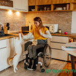 A women in a wheelchair and cat reaching up towards her in a kitchen