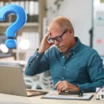 A man sitting at a desk holding his head, a laptop and bookcase and a Question mark