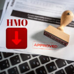 HMO texts above a red arrow pointing down in a red box, approved sign next to a stamp sitting on official papers on top of a keyboard