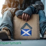 A homeless person sitting down holding a piece of cardboard with a Scottish flag