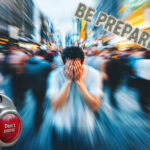 a man with his head in his hands in a large crowd, with a big red button saying don't panic.