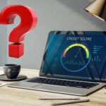 Laptop showing a credit score report on a desk with pens and a book, a red question mark sign above a coffee cup