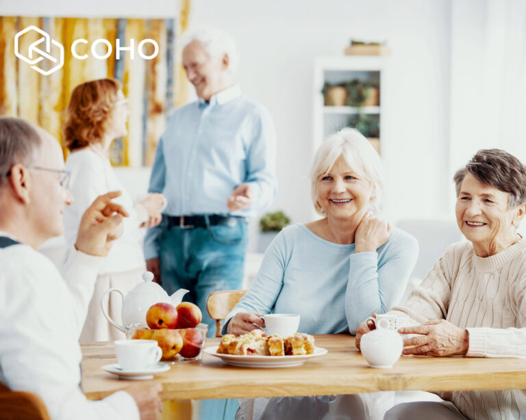 Older people sitting around a table drinking tea with cakes and fruit in a basket, people standing in the background
