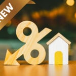 Wooden percentage symbol with downward arrow beside small house model, symbolizing reduced mortgage rates.