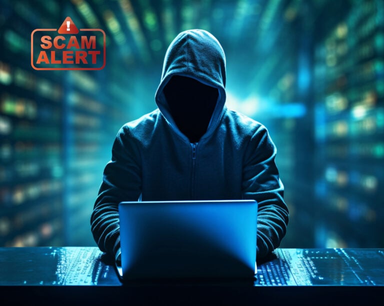 Hooded scammer at laptop with “Scam Alert” warning symbol.