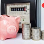 Piggy bank and coins beside electricity meter with question mark symbolizing rising energy costs.