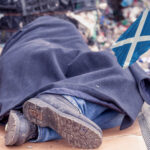 Person sleeping rough under a blanket with Scottish flag symbolizing housing crisis
