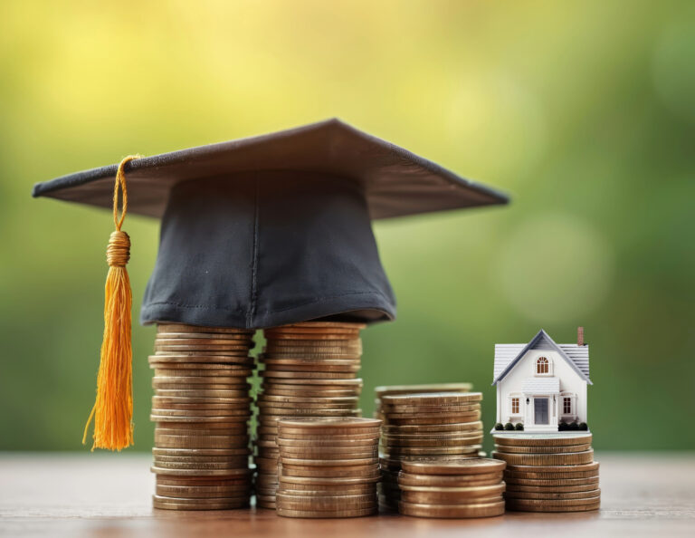 Stacks of coins with a graduation cap and miniature house symbolizing financial literacy and housing costs.
