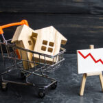 Wooden house models in a shopping cart beside a falling red trend line, symbolizing decline in housing sales.