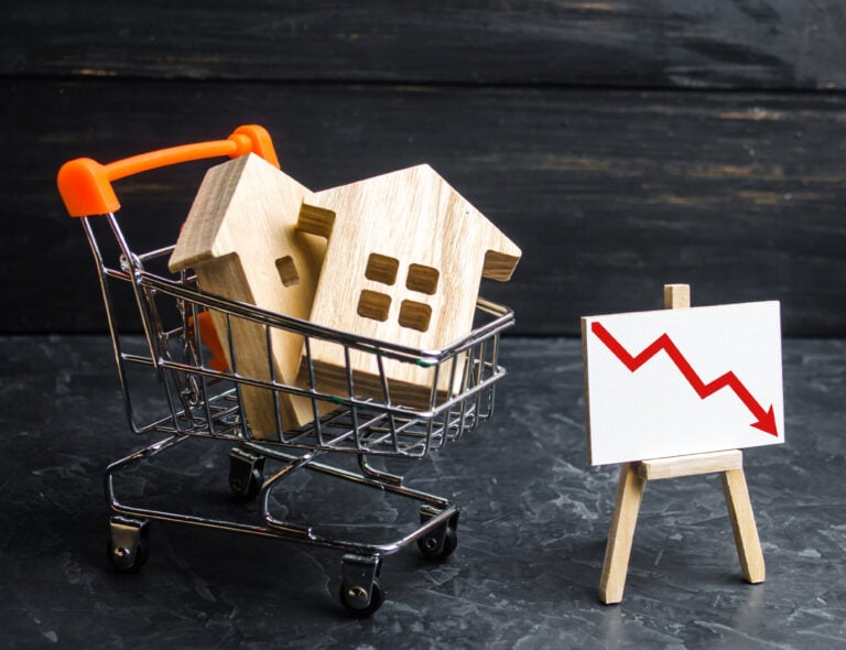 Wooden house models in a shopping cart beside a falling red trend line, symbolizing decline in housing sales.