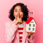 Woman signaling for silence while holding a model house with a "For Rent" sign.