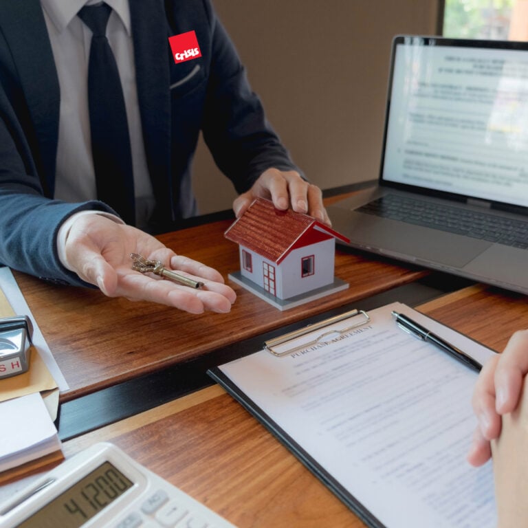 Estate agent handing over house keys beside model home and contract during property purchase meeting.