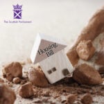 Wooden house model labelled “Housing Bill” leaning on rocks, representing Scotland’s new housing legislation.