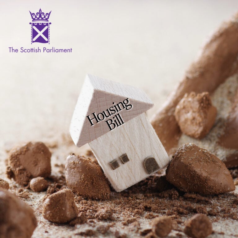 Wooden house model labelled “Housing Bill” leaning on rocks, representing Scotland’s new housing legislation.