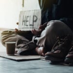 Person sitting on the ground holding a cardboard sign reading “HELP” to highlight homelessness crisis