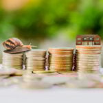 Slow growth in house prices illustrated by a snail on coin stacks beside a model house.