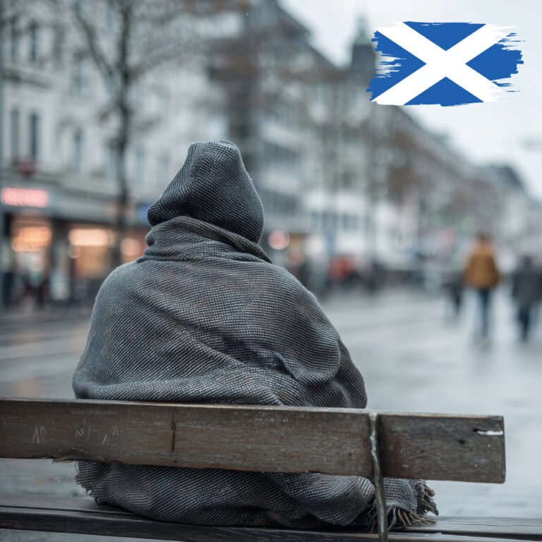 Person wrapped in a blanket sitting alone on a bench during cold, wet weather in Scotland