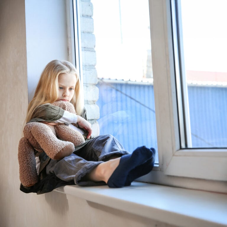 Child sitting on a windowsill hugging a teddy bear, reflecting the impact of homelessness on children
