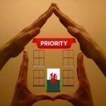 Hands forming a house shape around a Welsh flag door beneath a “Priority” banner, symbolising housing policy in Wales