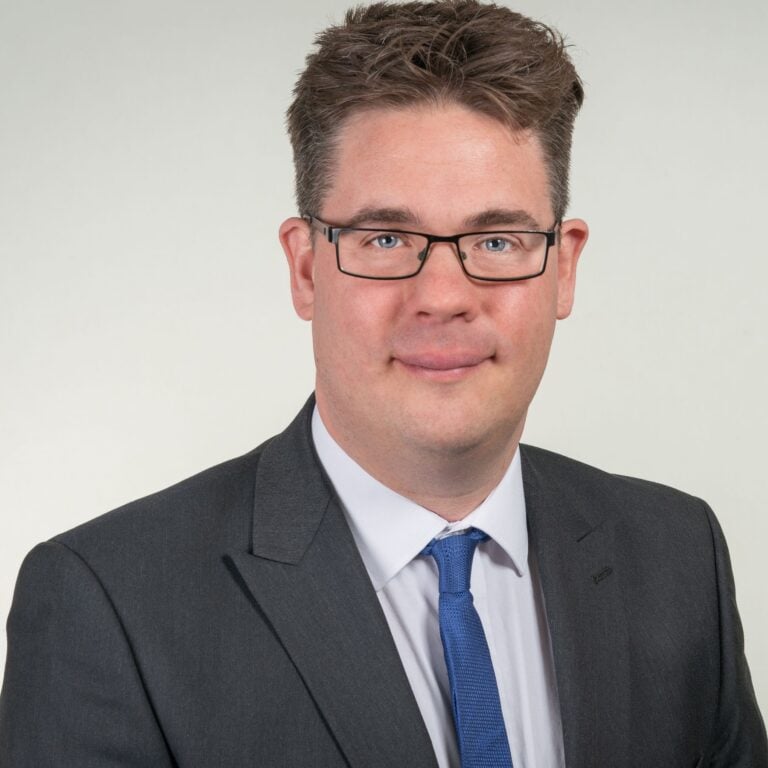 Daniel Smith of Gardner Leader wearing a grey suit, white shirt and blue tie, smiling in a professional headshot against a neutral background.