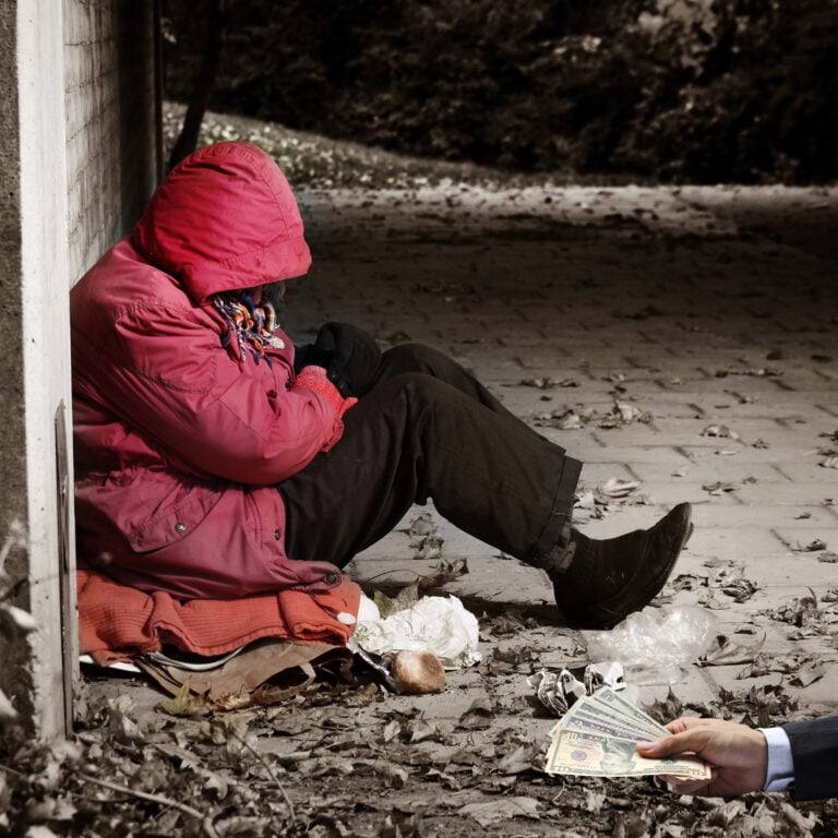 Person in a red coat sitting on pavement receiving money, illustrating homelessness and housing support efforts